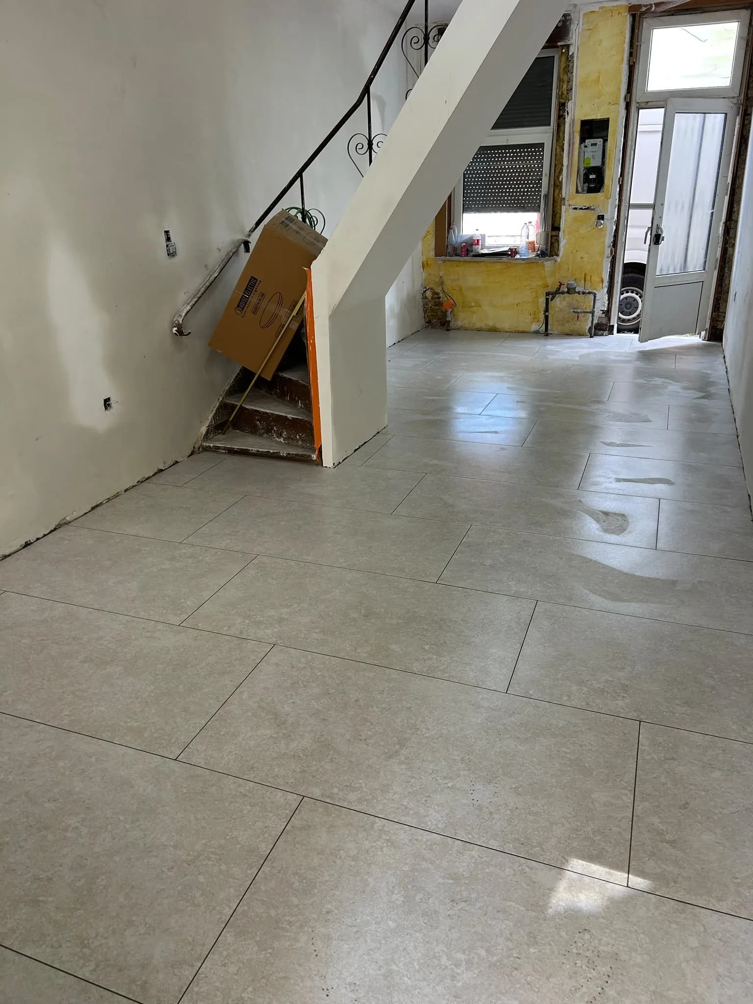 Pièce de vie carrelée en grand format clair par Km pose, escalier à gauche et porte d'entrée au fond